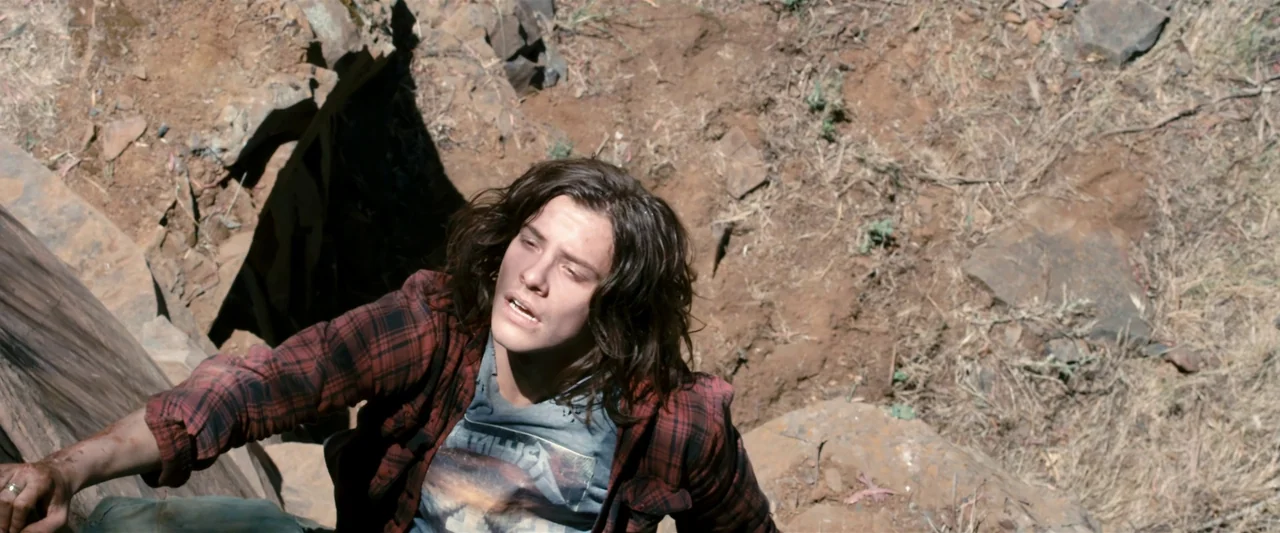The Loved Ones (2009) movie still: medium — A person climbs a steep, rugged earthen slope covered in rocks and sparse dry vegetation. The subject…