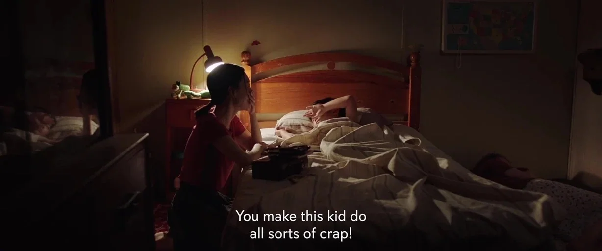 Minari (2020) movie still: medium — A dimly lit room contains a wooden headboard and a bedside table with a lamp. A woman sits on the edge of a bed…