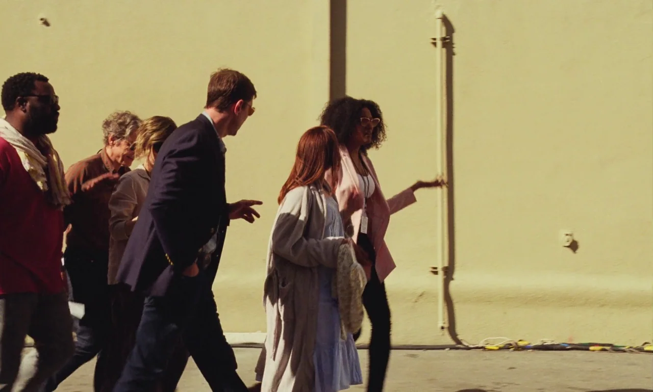 Marriage Story movie still: fullbody — A group of five people walks along a smooth beige exterior wall in daylight. The individuals are moving from…
