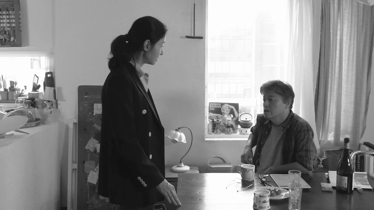 Walk Up (2022) movie still: medium — A modest room features a table set with drinks and miscellaneous small household items. A woman in a black…