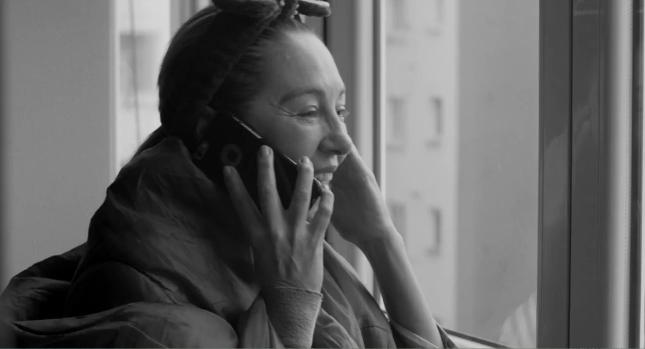 El Planeta (2021) movie still: closeup — A woman stands near a window inside a room with exterior light visible through glass. She is talking on a…