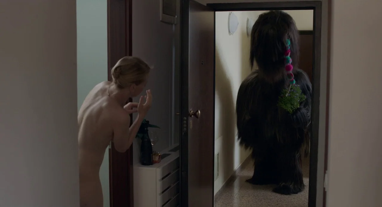 Toni Erdmann (2016) movie still: medium — An apartment hallway features a tiled floor and an open door frame. A nude woman peers out while a large,…