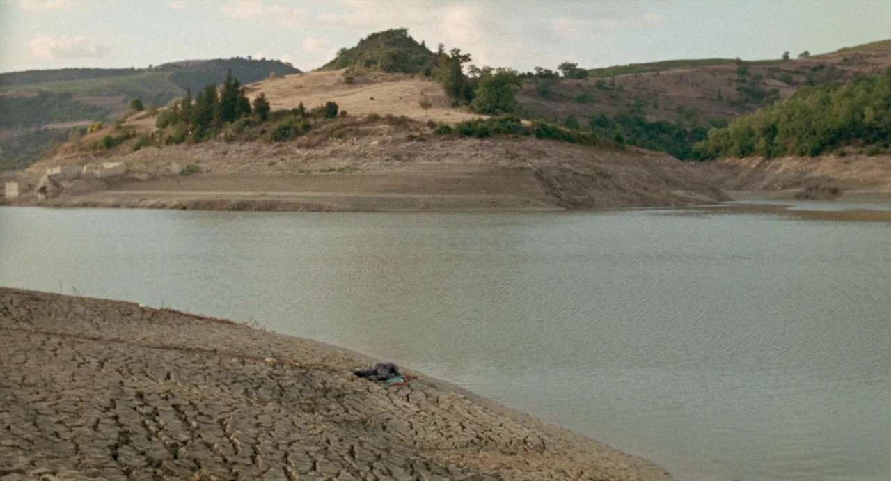 The Hellbenders (1967) movie still: wide — A dry, cracked shoreline leads into a large, murky lake surrounded by rolling hills. A small, indistinct…