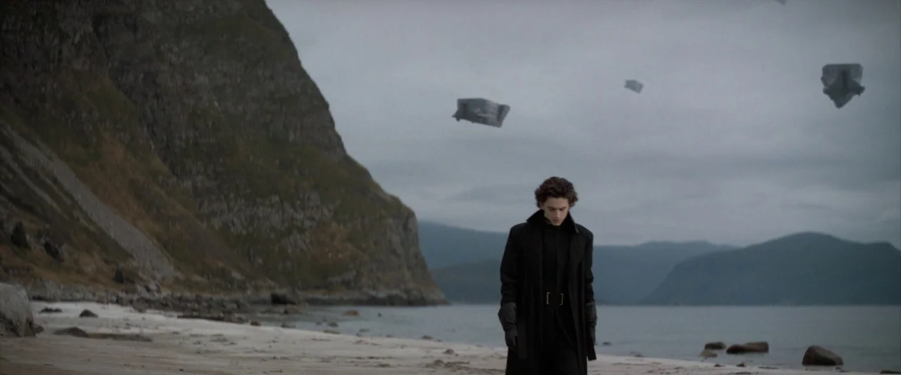 Dune (2021) movie still: fullbody — A desolate rocky shoreline stretches beside calm water under a heavy grey cloudy sky. A young man in a long dark…