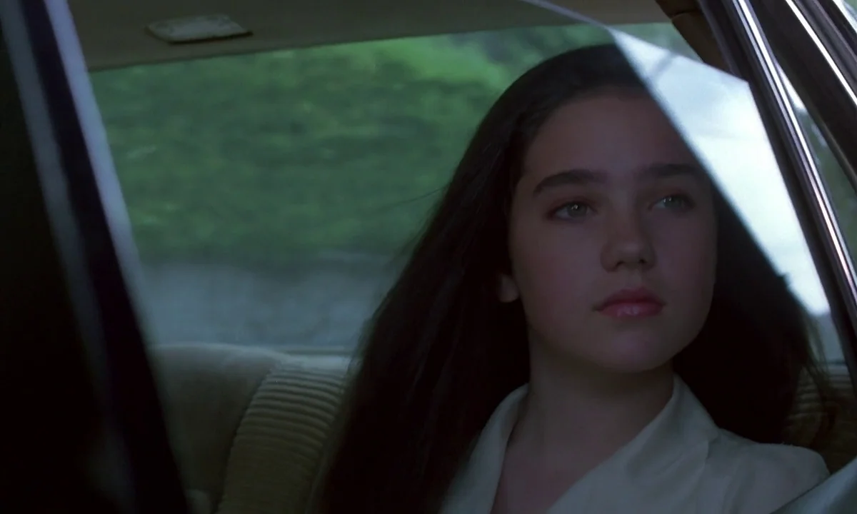 Phenomena (1985) movie still: closeup — The interior of a moving car showing the back seat upholstery. A young woman with long dark hair looks out…