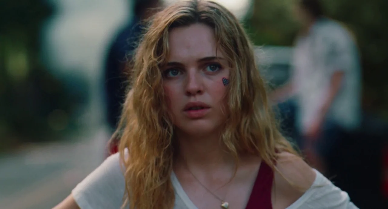 The Giant (2019) movie still: closeup — A woman stands in an outdoor area with blurred figures visible in the distance. She looks toward the camera…