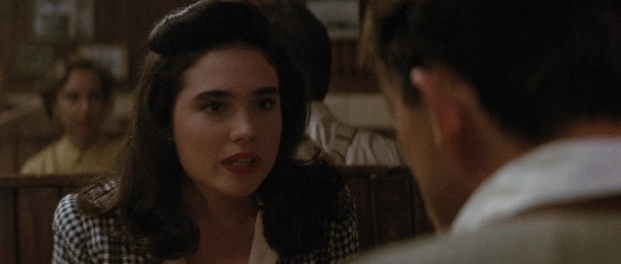 The Rocketeer (1991) movie still: medium — A dimly lit restaurant booth with dark wooden paneled walls in the background. A young woman speaks to a…