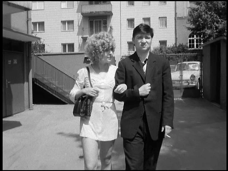Katzelmacher (1969) movie still: fullbody — A paved courtyard sits before a multi-story apartment building with balconies. A man and a woman walk…