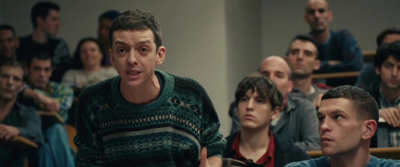 BPM (Beats Per Minute) (2017) movie still: medium — A crowded tiered lecture hall features rows of students sitting at wooden desks. A man in a…