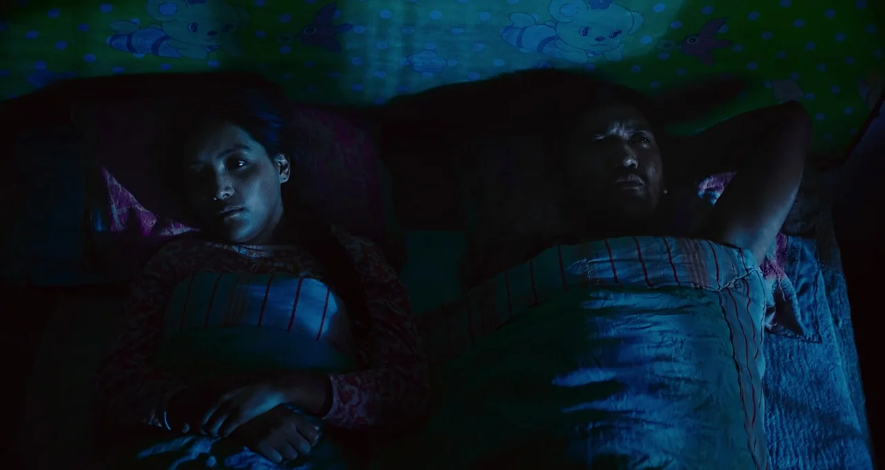 Balloon (2019) movie still: medium — A dark, dimly lit space with patterned green bedding visible above the two people. A man and a woman lie…