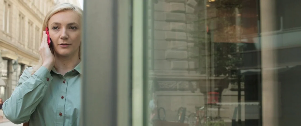 On Body and Soul (2017) movie still: medium — A woman stands on a city sidewalk next to a large glass window pane. She holds a bright red mobile…