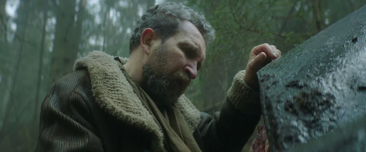 Vesper (2022) movie still: medium — A man stands in a dense, overcast forest near a wet, dark, vertical object. The man reaches out with his hand to…