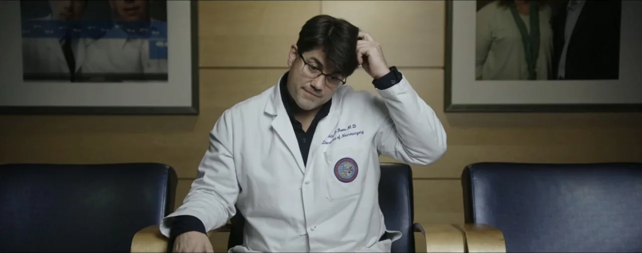 Hunting for Hedonia (2019) movie still: medium — A man in a medical coat sits in a waiting room with wood paneling. The man looks down while…