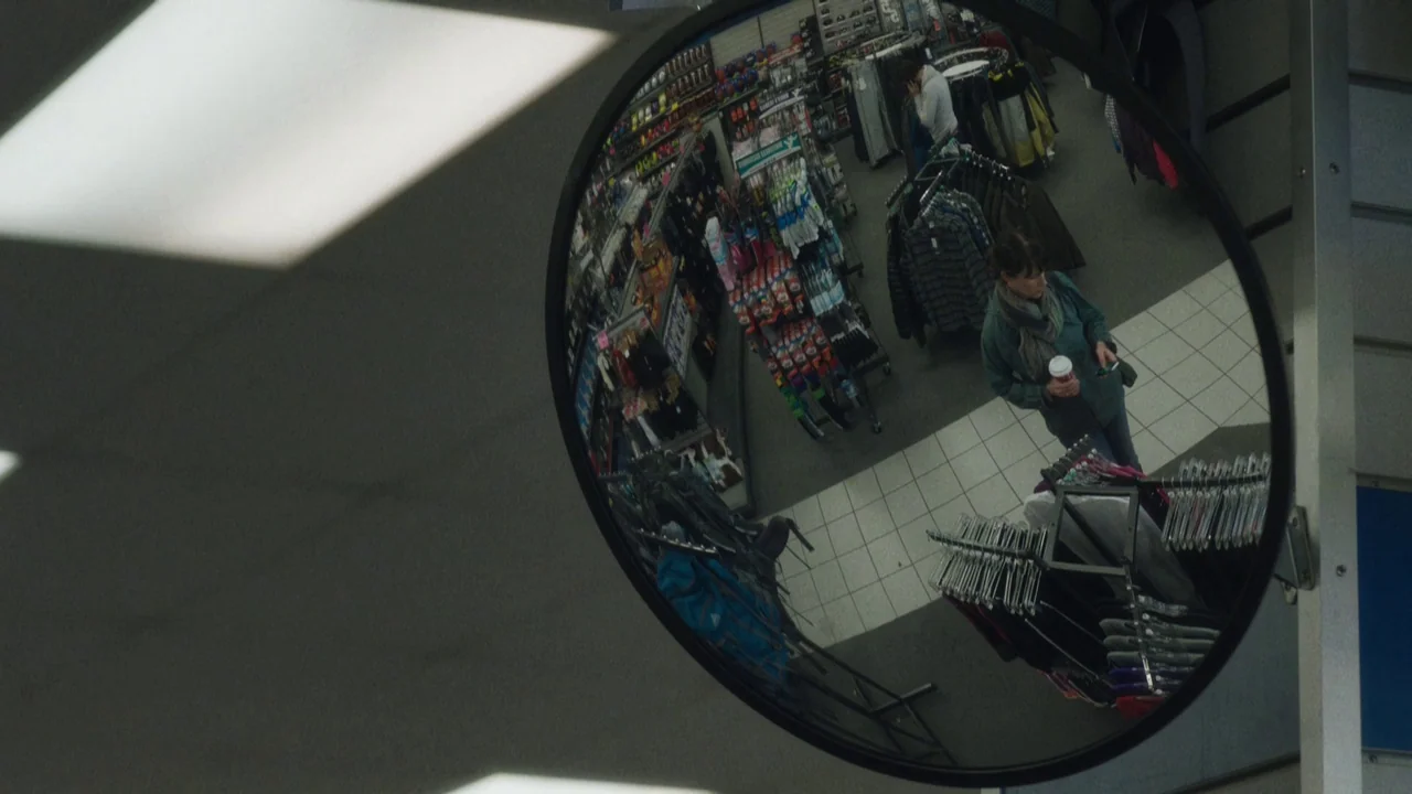 Night Moves (2013) movie still: medium — A view of a retail aisle through a circular convex safety mirror. A woman stands in the aisle holding a…