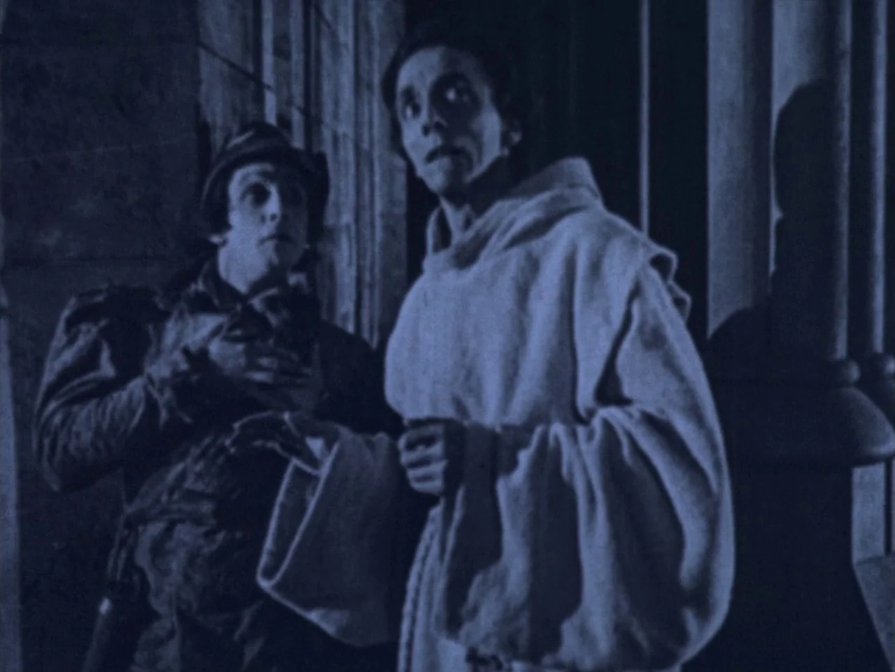 The Hunchback of Notre Dame (1923) movie still: medium — A stone corridor with tall pillars illuminated by high-contrast blue light. Two men dressed…