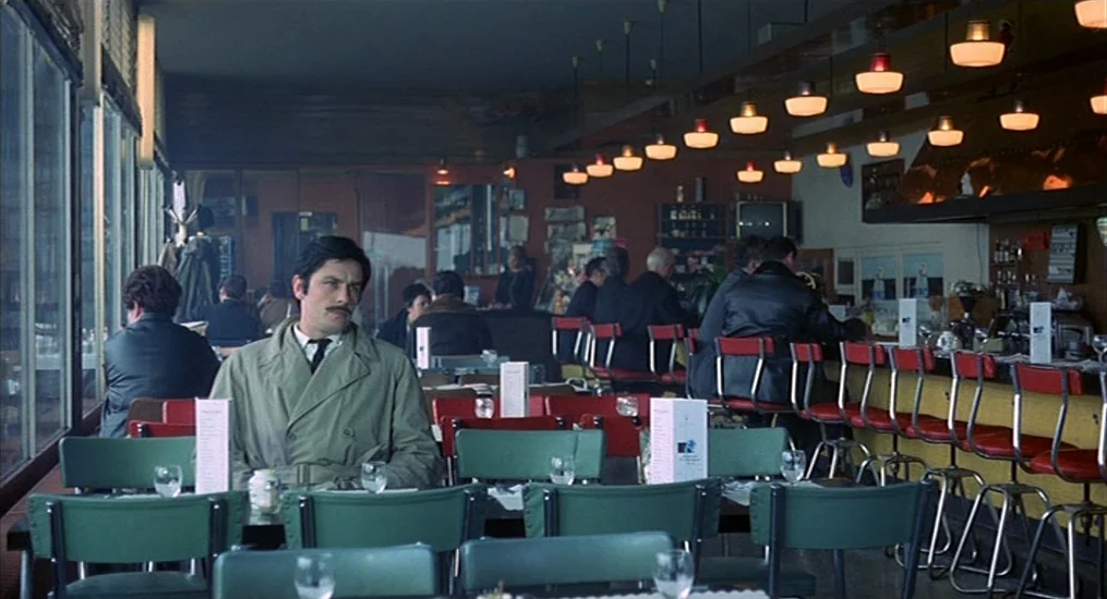 Le Cercle Rouge (1970) movie still: medium — A mid-century diner with rows of green chairs and red-seated bar stools. A man sits at a foreground…