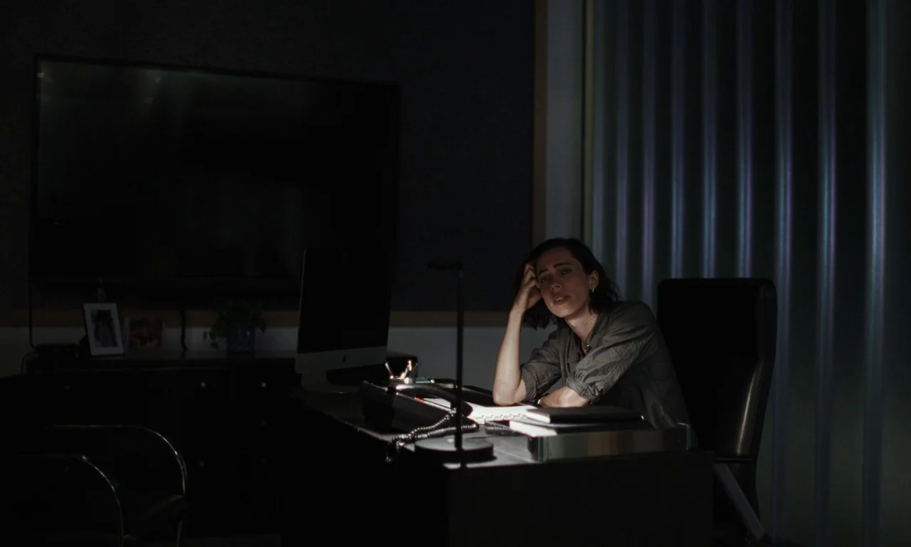 Resurrection (2022) movie still: medium — A dark, dimly lit professional workspace features a black desk and wall-mounted screen. A woman sits…