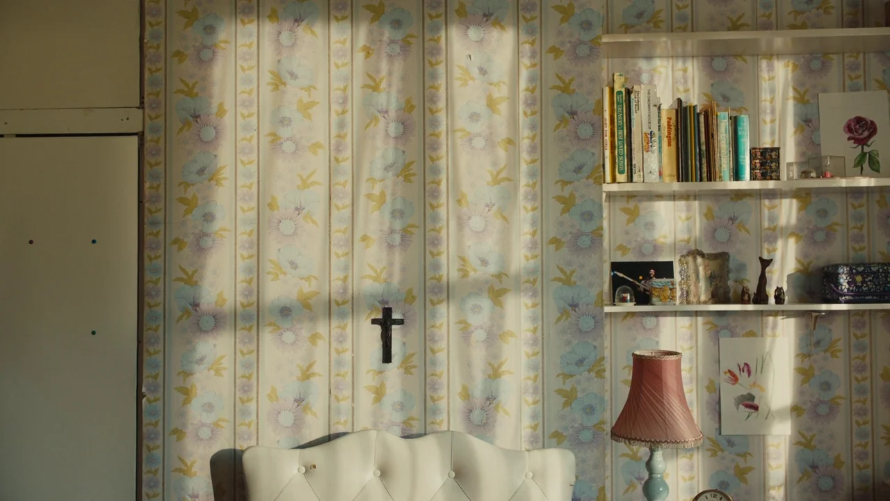 Lover’s Rock (2020) movie still: wide — A wall covered in vintage floral wallpaper features built-in shelves containing books and small ornaments. A…