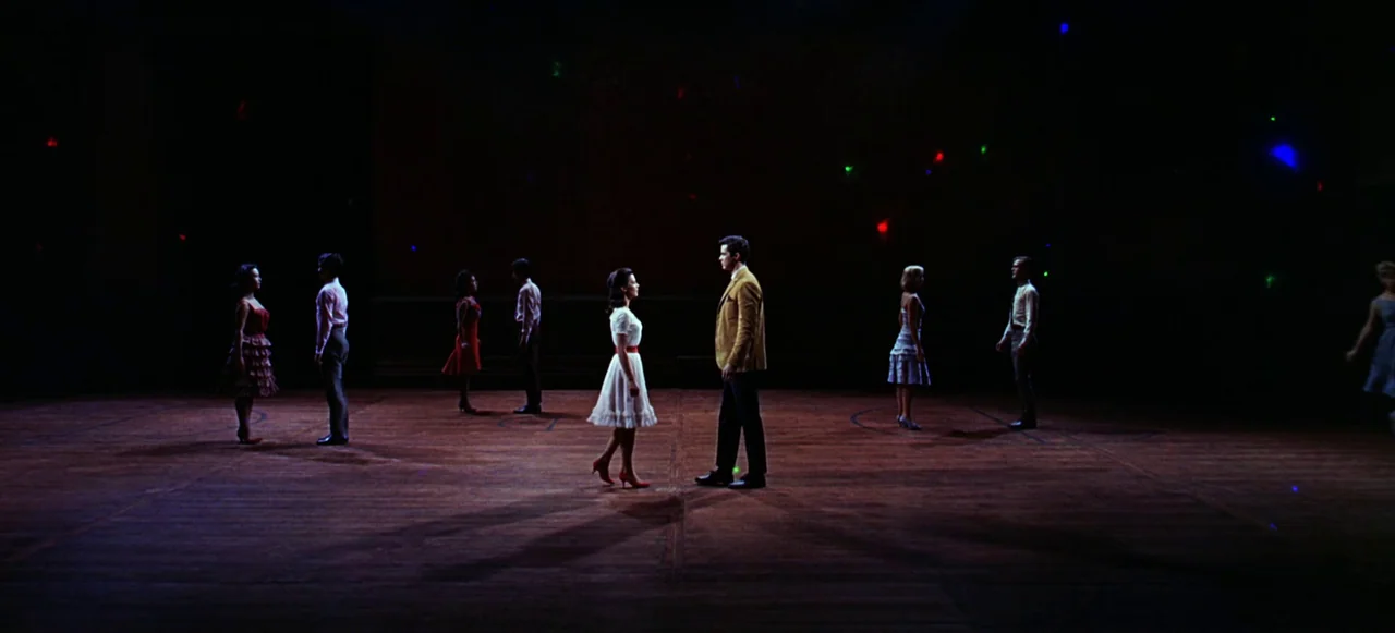 West Side Story (1961) movie still: wide — A spacious high school gymnasium floor features multiple couples standing apart in the shadows. Each pair…