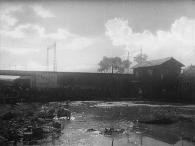 Drunken Angel (1948) movie still: establishing — A desolate, mud-filled lot contains scattered debris and a distant, weathered wooden house. A long…