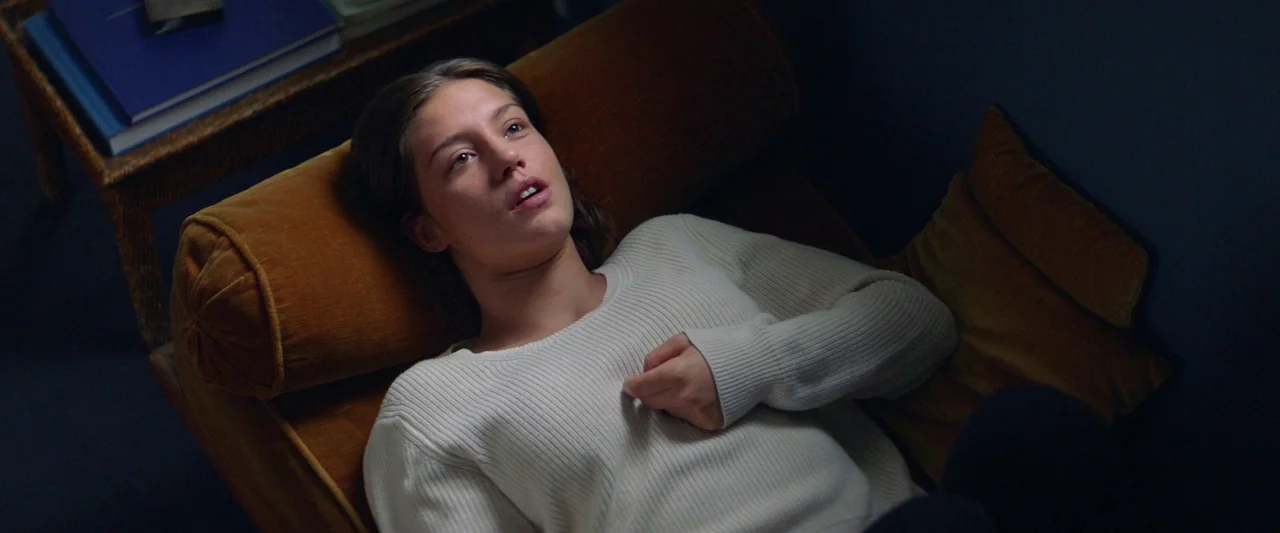 Sibyl (2019) movie still: medium — A woman rests on a deep amber velvet chair inside a dimly lit room. She looks upward with an expression of quiet…