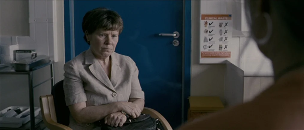 Another Year (2010) movie still: medium — A sterile medical room features a blue door and a clinical waste sign. An older woman sits in a chair…