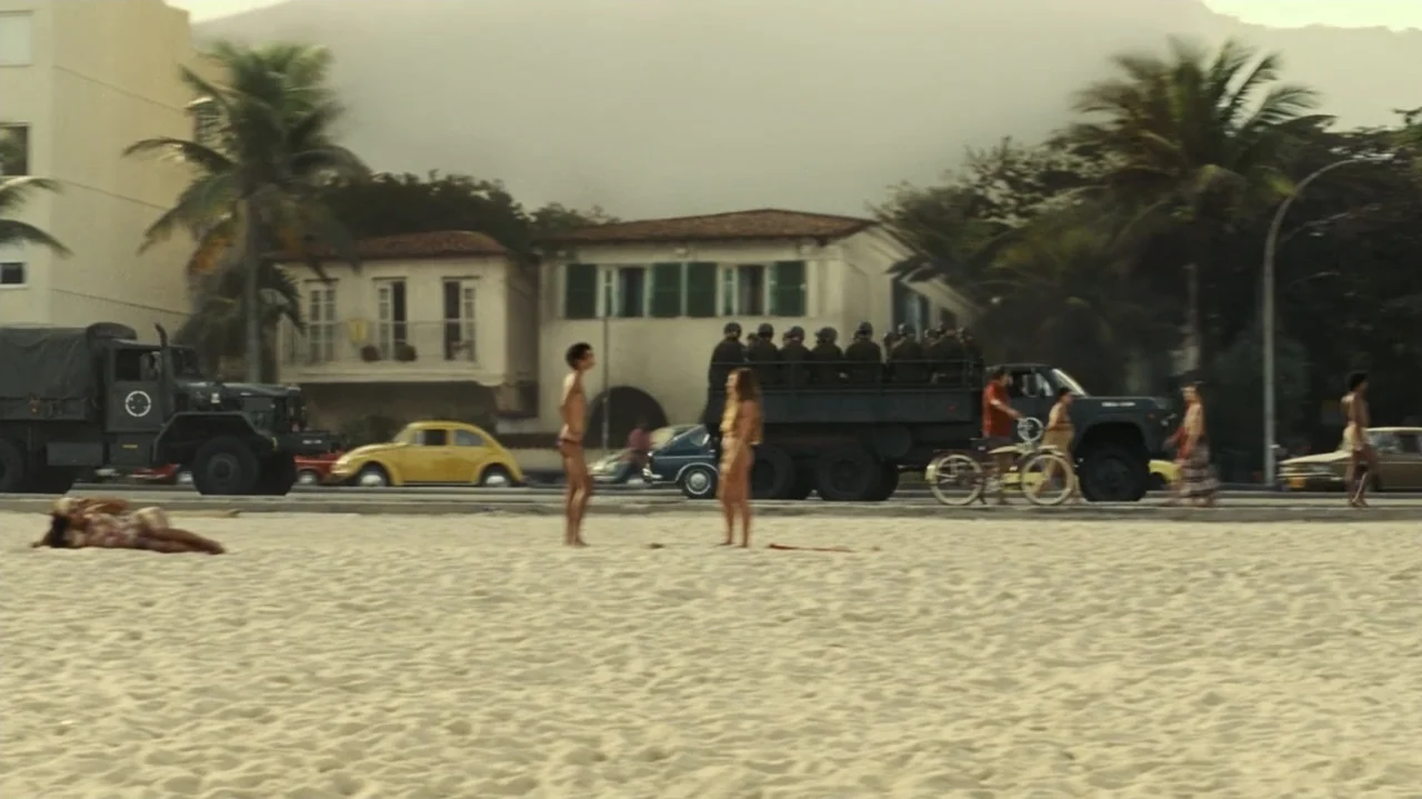 I'm Still Here (2024) movie still: wide — A sandy beach stretches out before a road lined with buildings and palm trees. A military truck carries…