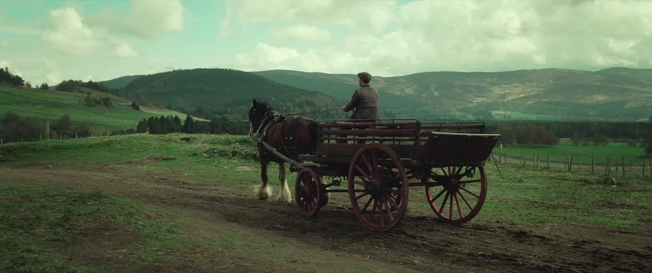 Sunset Song (2015) movie still: wide — A rural dirt path winds through rolling green hills under an overcast sky. A man in a tweed jacket drives a…