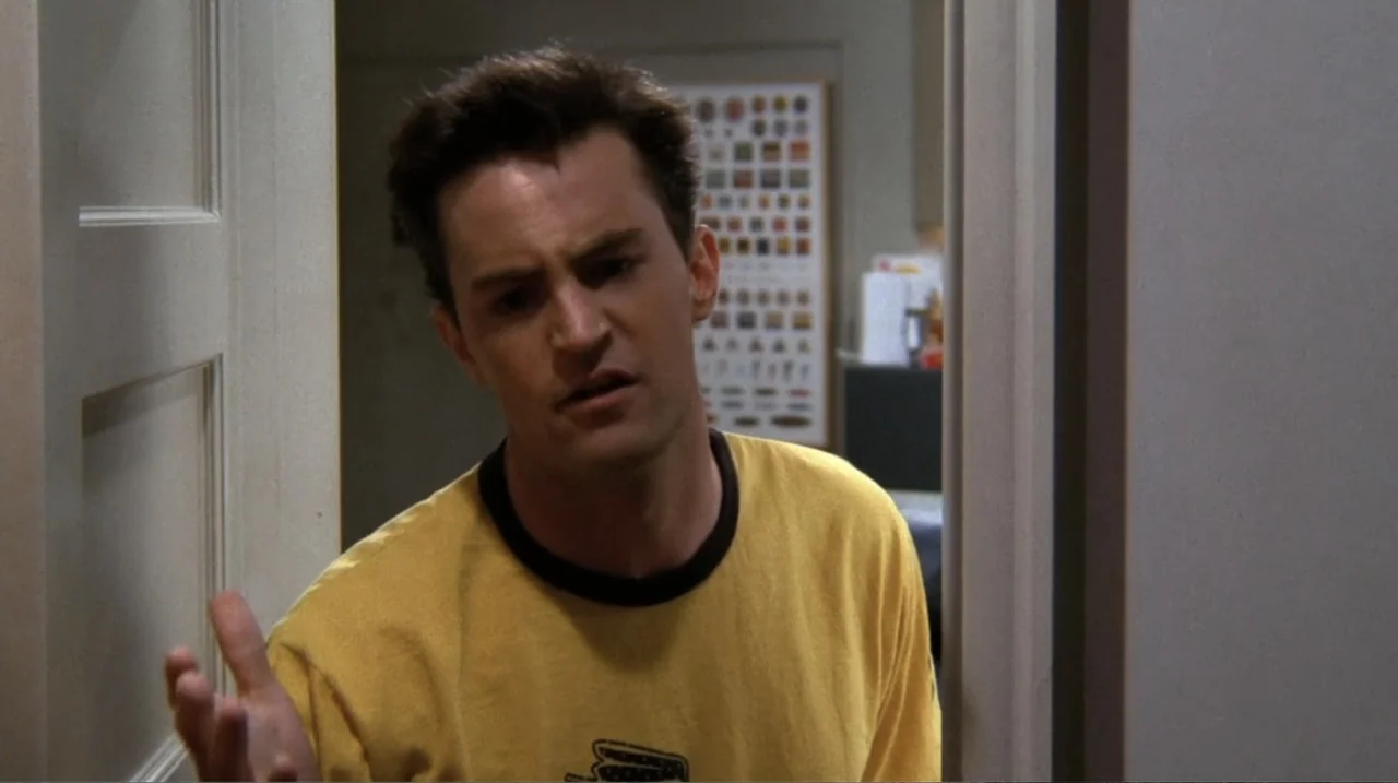 Friends: The Reunion (2021) movie still: medium — A man stands in an apartment doorway with a blurred poster on the back wall. The man wears a…