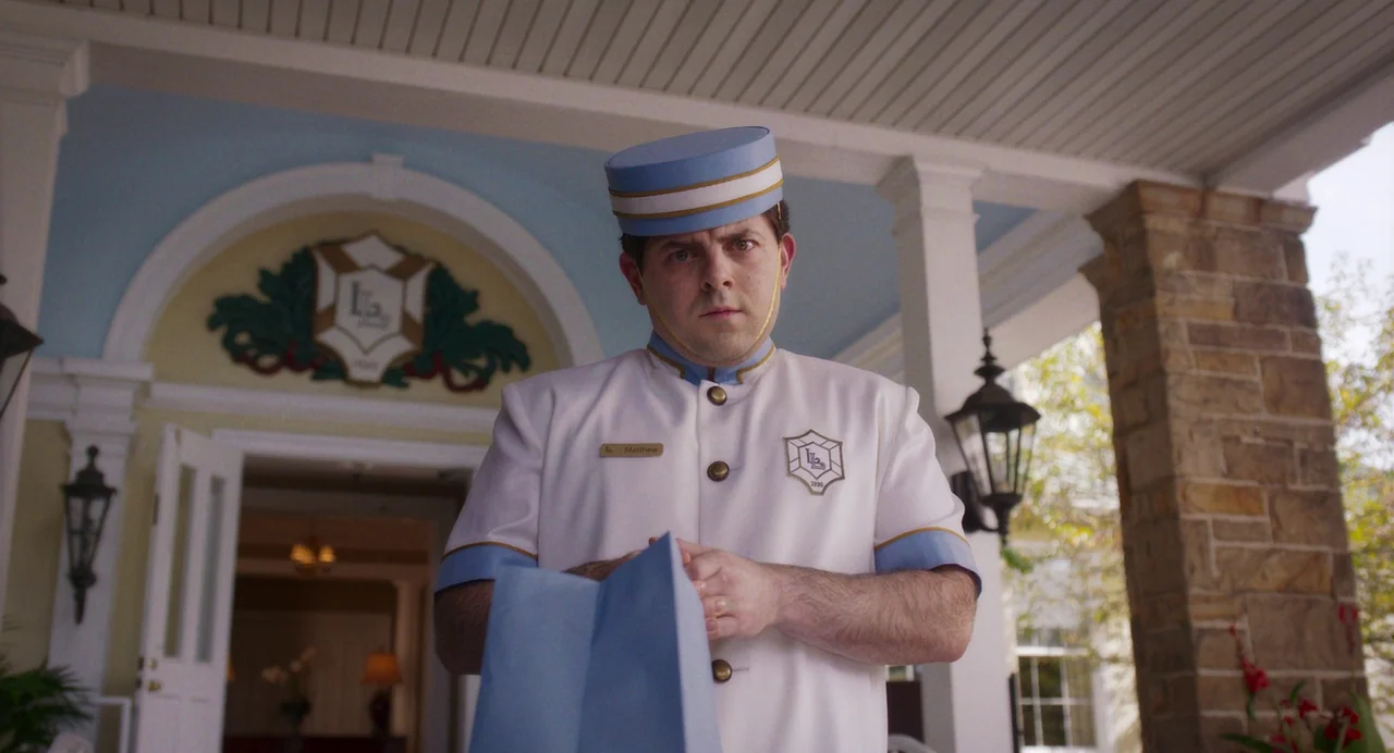 Drive-Away Dykes (2024) movie still: medium — A hotel entrance features a light blue arched doorway with a white stone facade. A male bellhop in a…