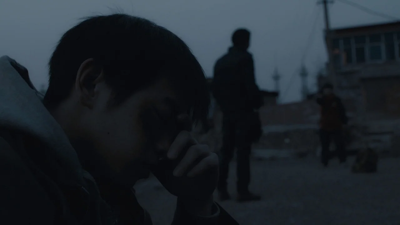 An Elephant Sitting Still (2018) movie still: closeup — A dark outdoor space with uneven dirt ground and a blurry building in back. A young man sits…