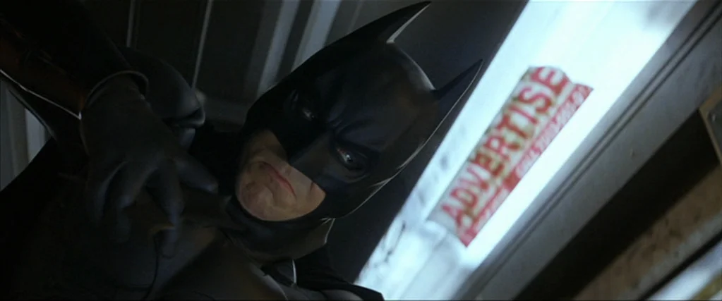 Batman Begins (2005) movie still: closeup — A dimly lit interior of a subway car featuring an advertisement on the wall. Batman looks down, his face…