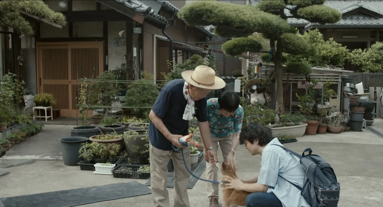 Ramen Shop (2018) movie still: fullbody — A residential courtyard filled with various potted plants and a pruned bonsai tree. Two elderly people and…