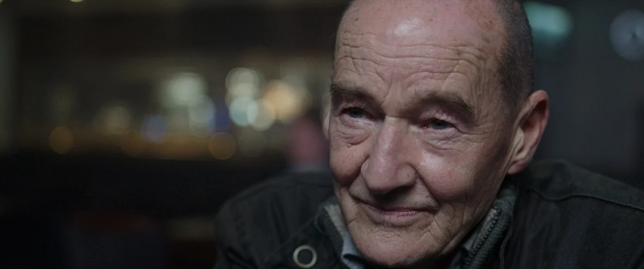 Bull (2021) movie still: closeup — An elderly man is positioned in an indoor space at night. The background features bokeh highlights from distant…
