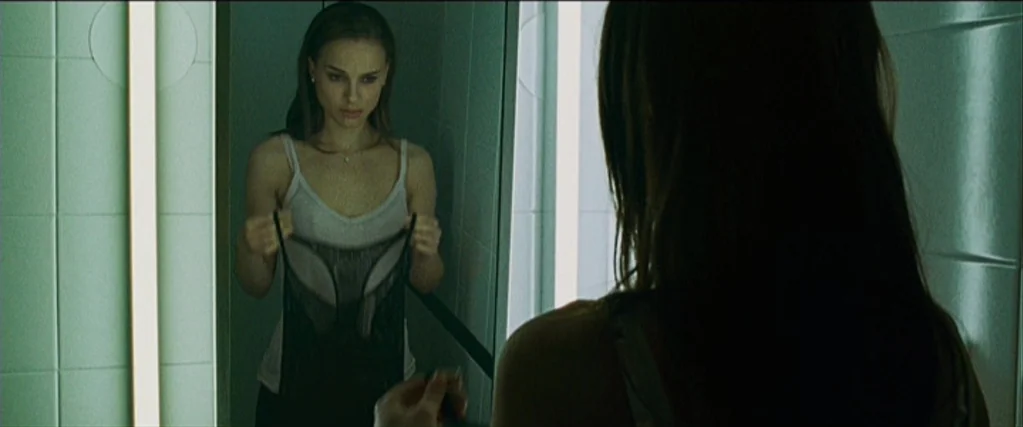 Black Swan (2010) movie still: medium — A woman stands before a large mirror inside a white-tiled bathroom. She holds a piece of black fabric in her…