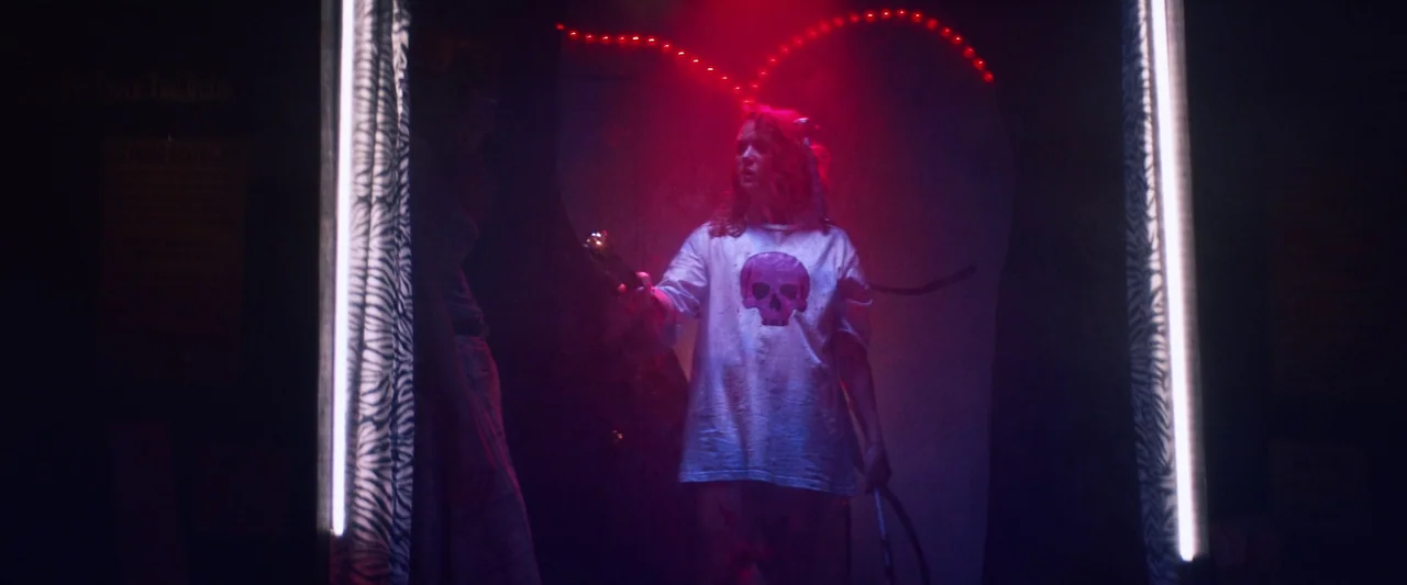 Revealer (2022) movie still: medium — A dark booth interior illuminated by a red neon heart shape on the back wall. Two young women stand in the…