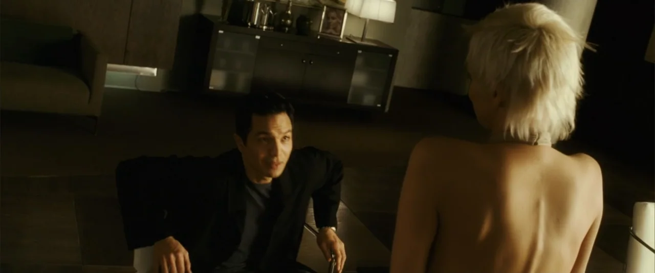 Catwoman (2004) movie still: medium — A modern, dimly lit luxury apartment features dark furniture and warm ambient lighting. A man sits facing a…