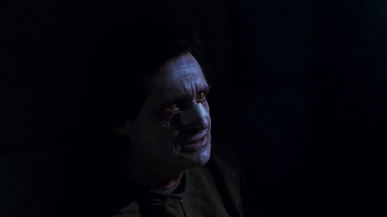 The Exorcist 3 (1990) movie still: closeup — A man with yellow eyes sits in a dark, shadow-filled environment. He has a bandage across his nose and…
