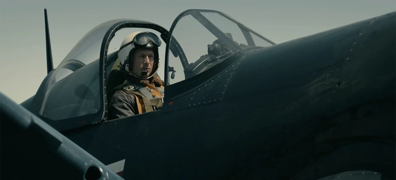 Devotion (2022) movie still: medium — A dark blue vintage propeller fighter plane sits under a clear sky. A pilot wearing a flight helmet and…