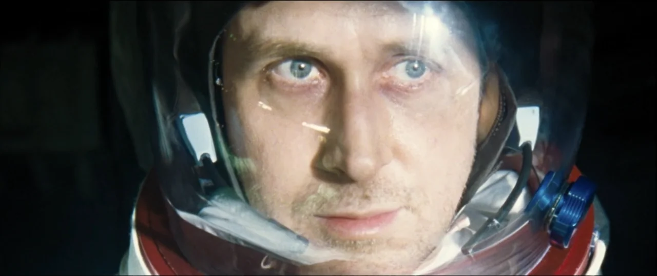 First Man (2018) movie still: closeup — A close view inside a pressurized space suit helmet shows a man's face. The clear visor reflects light while…