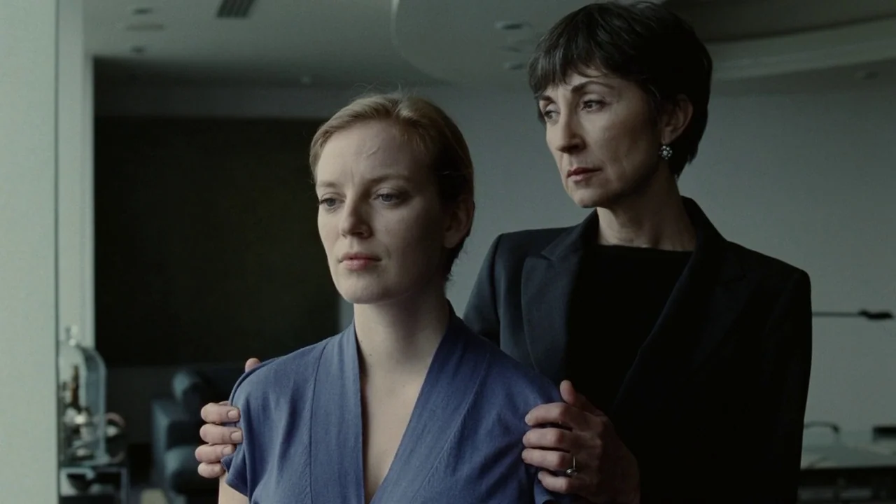 Splice (2009) movie still: medium — A spacious room with minimalist modern decor features large glass windows overlooking an outdoor area. An older…