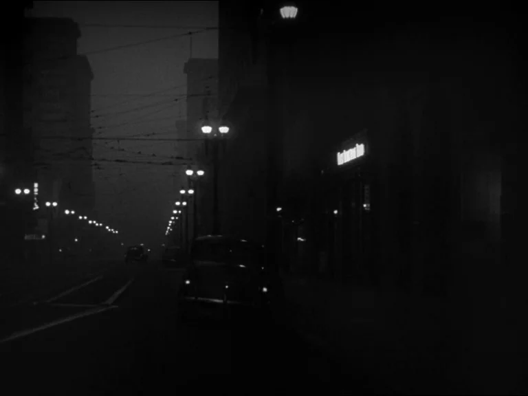 Double Indemnity (1944) movie still: establishing — A dark, deserted city street at night is lined with glowing overhead streetlights. An antique…