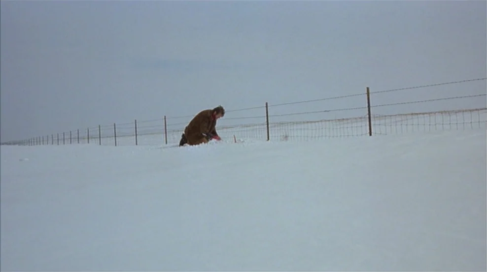 Fargo (1996) movie still: wide — A vast, flat, snow-covered landscape stretches under a pale, overcast sky. A single man in a brown coat kneels by a…