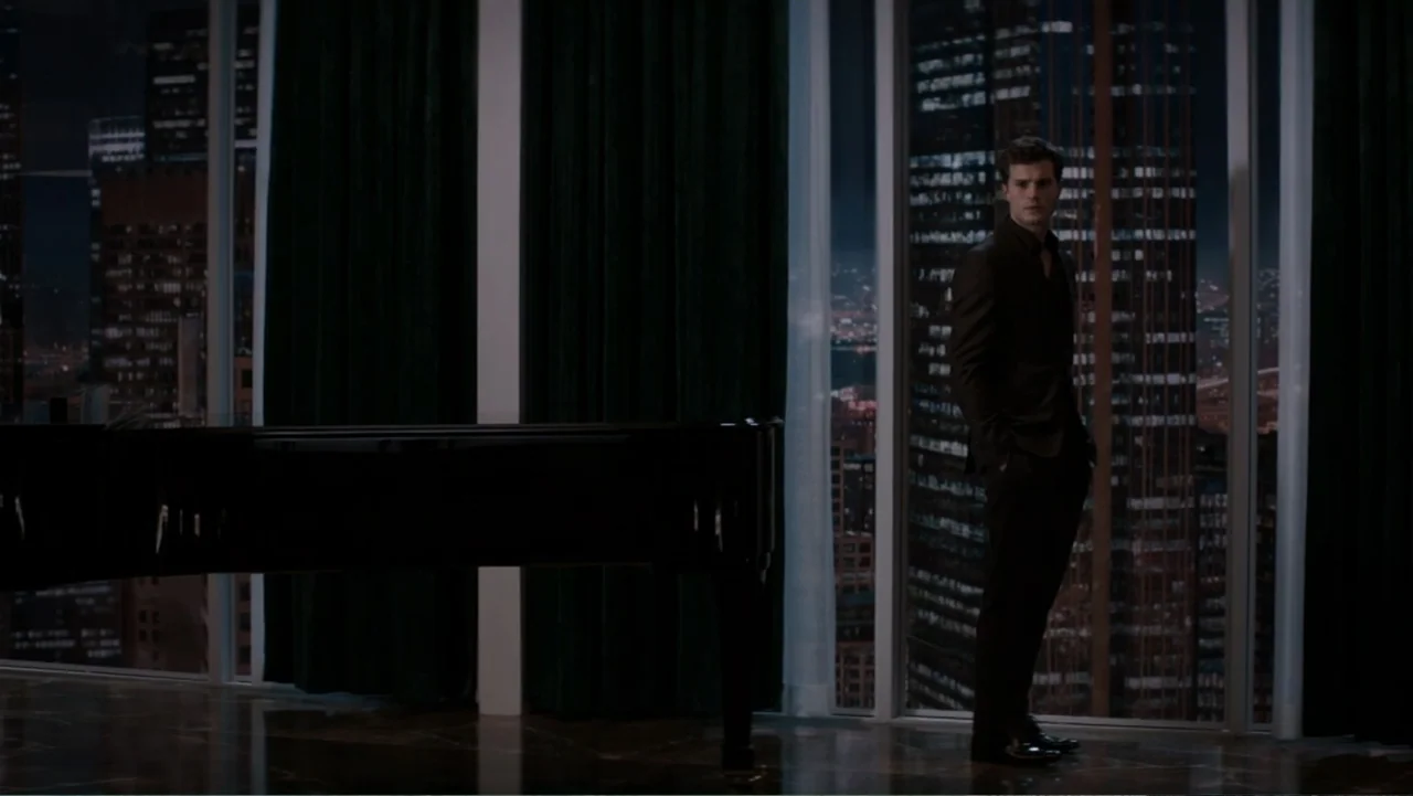 Fifty Shades of Grey (2015) movie still: fullbody — A high-rise interior features dark curtains and a glossy floor near a large window. A man in a…