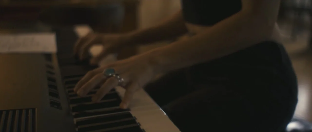 Night of the Missing (2023) movie still: closeup — A dimly lit room contains a digital piano positioned in front of a person. A person with a silver…
