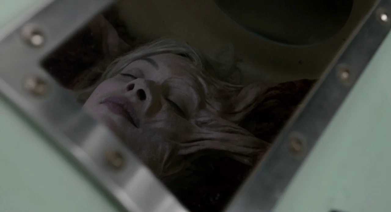 Antiviral (2012) movie still: closeup — A woman's head rests inside a metallic containment unit with a viewing window. She lies submerged and…