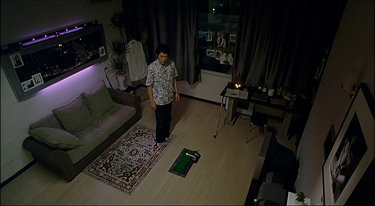 3-Iron (2004) movie still: wide — A modern apartment room features a patterned rug and a putting green mat. A man stands alone on the putting mat…