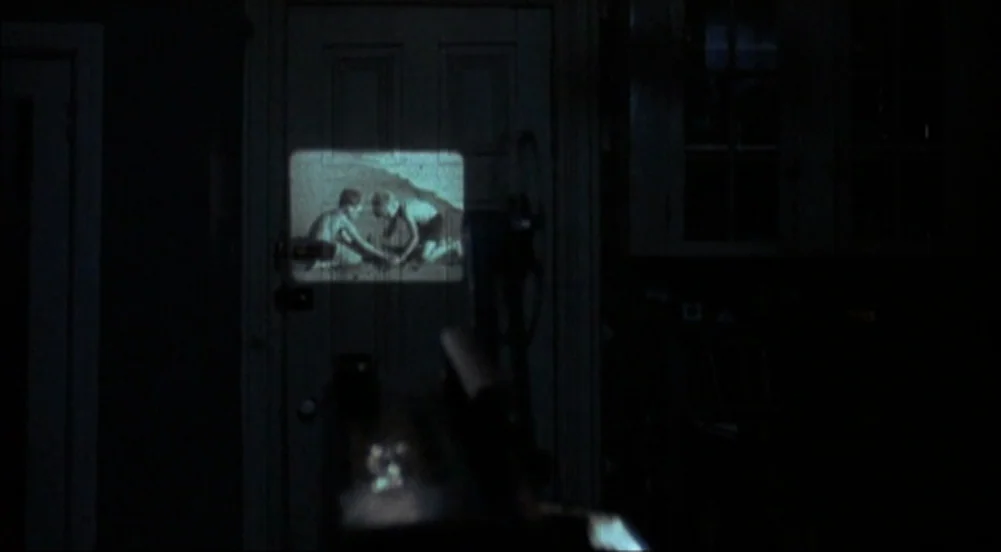 The King Of Marvin Gardens movie still: wide — A dark, dimly lit room contains a door in the center of the frame. A small home movie is projected…