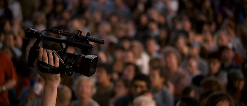 This Movie Is Broken (2010) movie still: medium — A dense crowd of people is blurred in the background of an outdoor space. An arm holds a…