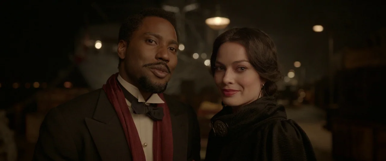 Amsterdam (2022) movie still: medium — A night scene at a harbor pier with blurred streetlights glowing in the background. A man in a tuxedo and a…